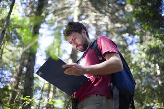 homme au t-shirt bordeau, portant un sac à dos, lisant un rapport dans la forêt