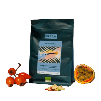 Un paquet de tisane Packshot_L_ensoleillée__1_-removebg-preview vert foncé, mettant en vedette Thé vert à la menthe, est astucieusement entouré d'églantier frais, d'un fruit de la passion tranché et de tranches de gingembre pour mettre en valeur son mélange d'ingrédients naturels.