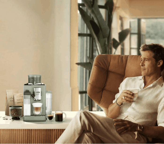 Une personne se détend dans un fauteuil confortable, une tasse de café à la main. À proximité, sur la table, se trouve une machine mobile Delonghi Rivelia-Green-Herobanner-mobile entourée d'un assortiment de boissons et de grains de café. De grandes fenêtres et une plante renforcent l'atmosphère chaleureuse et bien éclairée, créant le cadre idéal pour savourer votre boisson préférée.